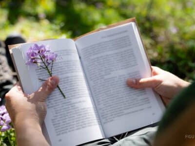 Manos sosteniendo un libro con un separador de flores en un bosque.