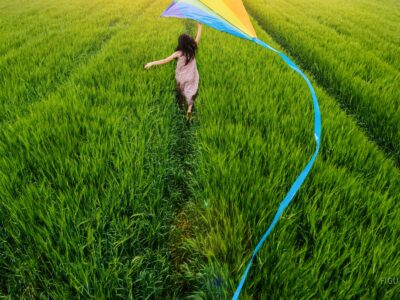 Niña corriendo con un papalote de colores en un campo de trigo verde.